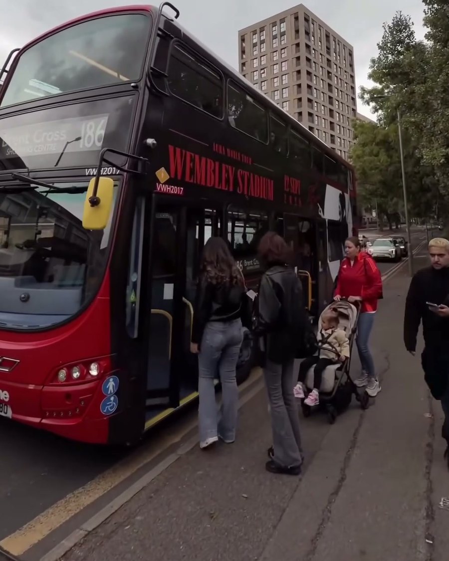 Passengers boarding the Aura World Tour wrapped bus at a stop, Wembley Stadium and 12 Sept 2026 above the doors
